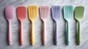 Seven pastel-colored kitchen spatulas, made of rubber or silicone, are arranged in a row on a marble surface, each showcasing a unique color and design.